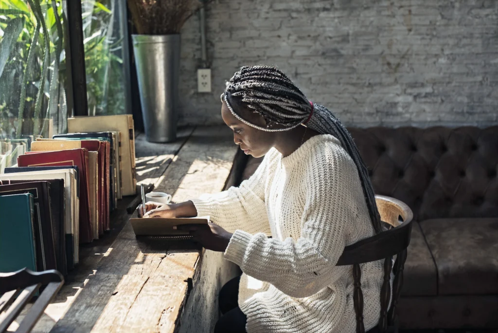 Femme noir avec des tresses qui écrit dans son journal assise à un bureau